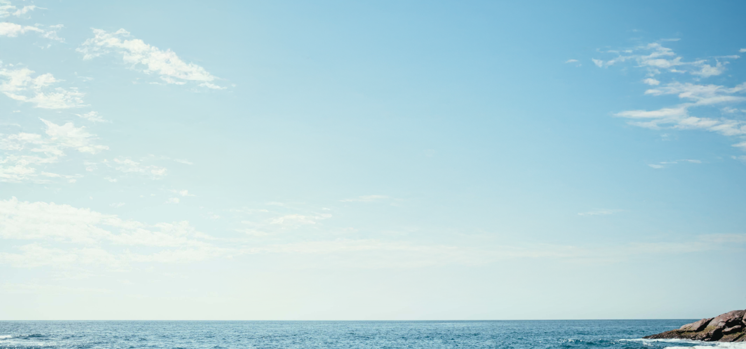 image of large blue sky with sparse, whispy clouds, a calm sea and the start of a rock formation peeking in from the lower right corner.
